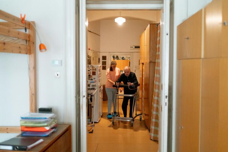 A caregiver assists an elderly man with a walker in a comfortable home environment.