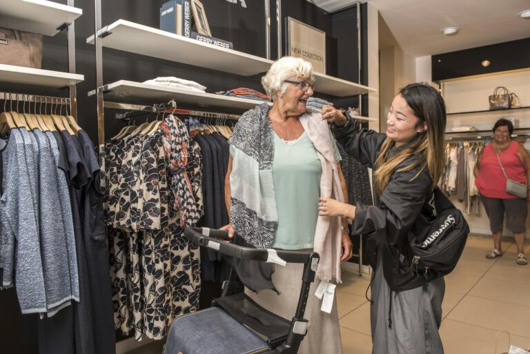 A senior woman is being helped by a caregiver while shopping in a clothing store. She uses a walker for mobility.